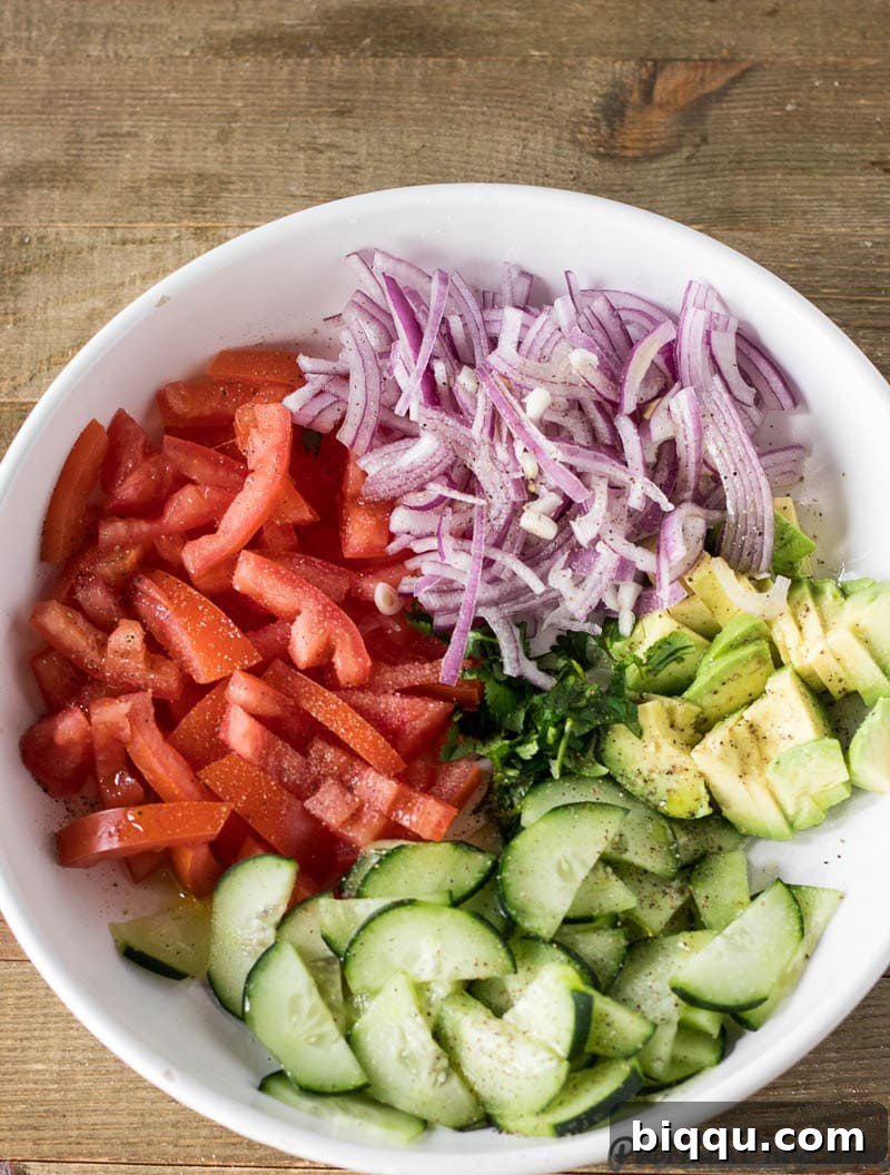 A close-up shot of a bowl of Simple Kenyan Salad, featuring diced tomatoes, red onions, cucumber, and avocado with black pepper specks.