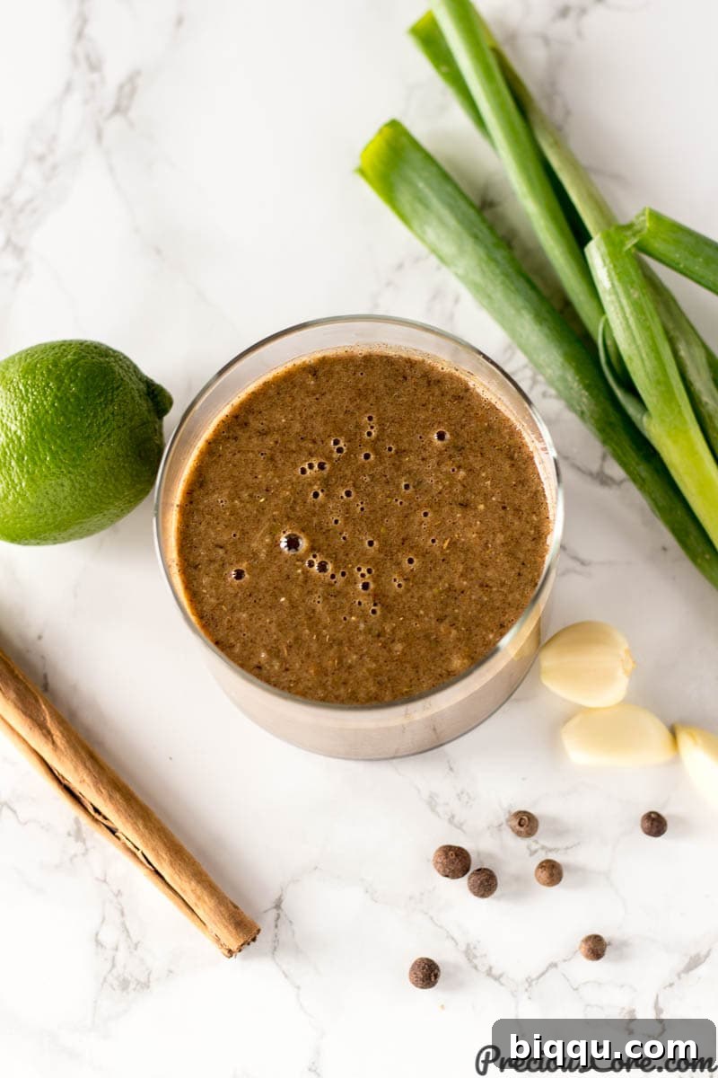 Glass jar of homemade Jamaican jerk seasoning surrounded by fresh ingredients like garlic, lime, and Scotch bonnet peppers on a wooden table.