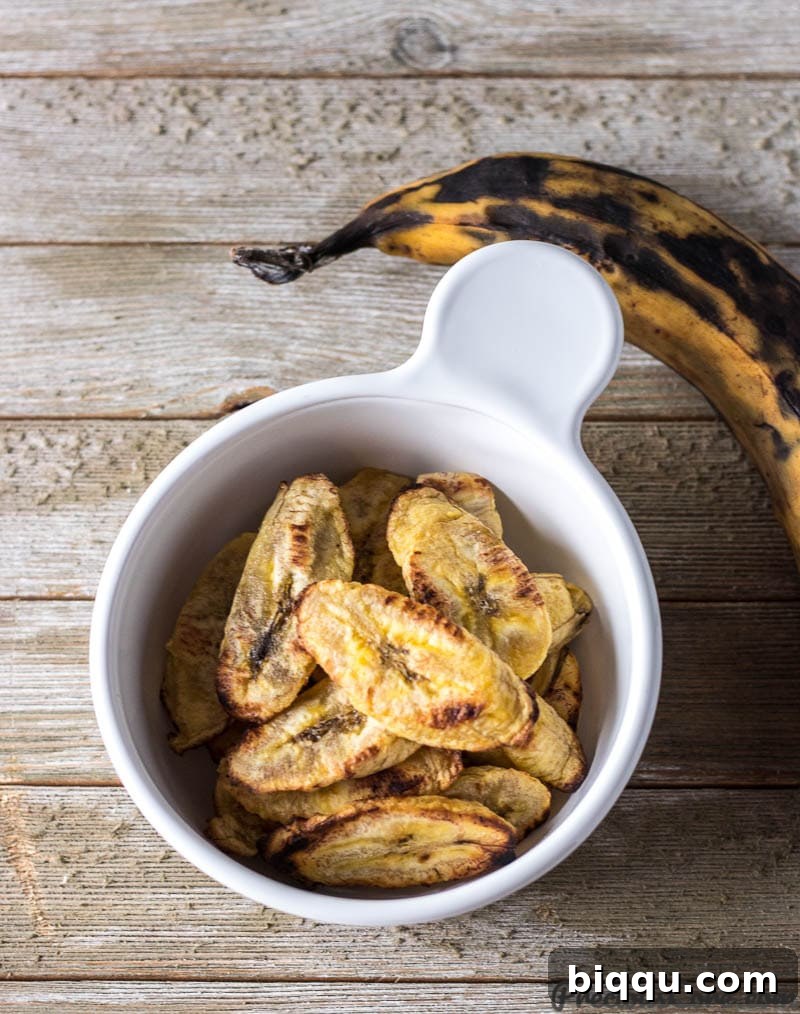 A bowl of freshly baked plantains with one whole uncooked ripe yellow plantain beside it, showcasing the ideal ripeness for baking