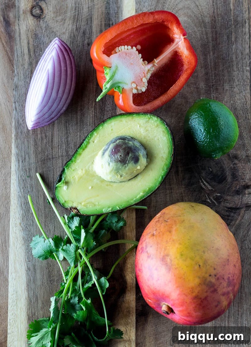 Fresh ingredients laid out on a wooden chopping board: vivid red onion, crisp red bell pepper, a juicy lime, creamy avocado, fragrant cilantro, and a perfectly ripe mango.