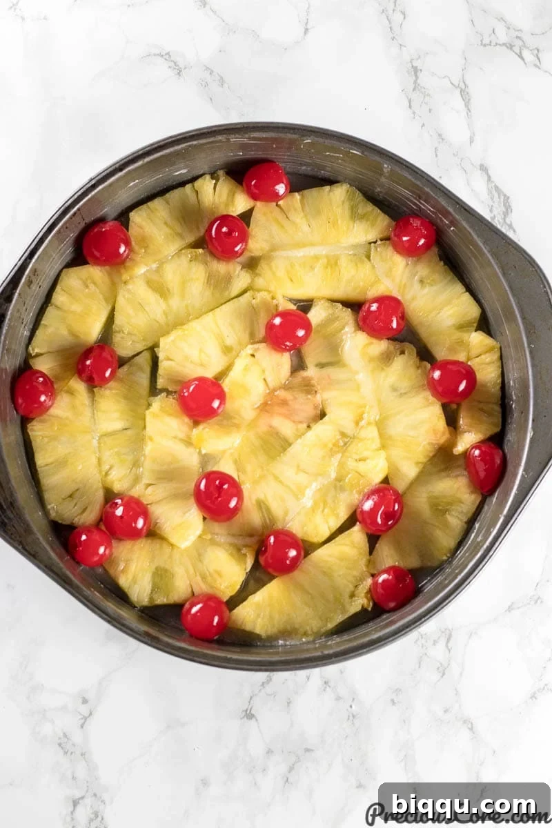 A baking pan meticulously layered with golden pineapple slices and bright red maraschino cherries, forming the beautiful base of the Pineapple Upside Down Cake before the batter is added.