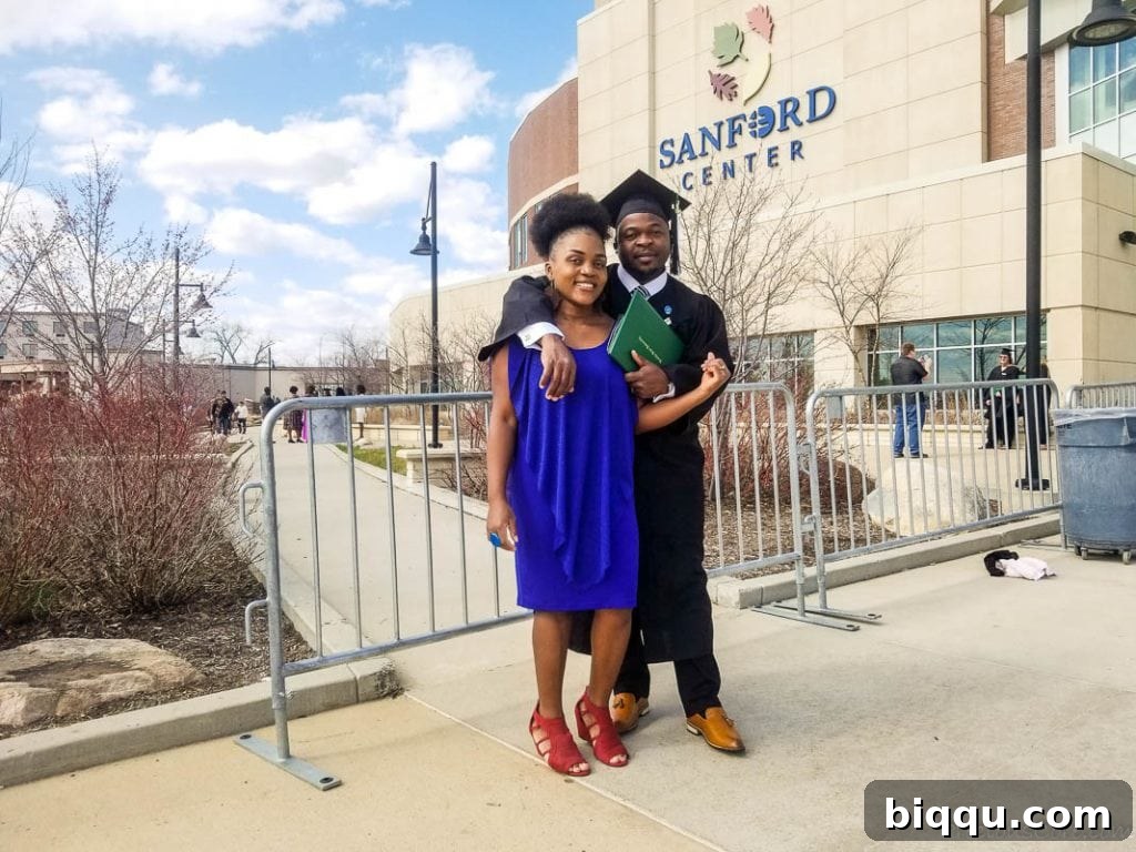 A proud man in graduation attire stands beside a beaming woman in a blue dress, in front of the Sanford Center.