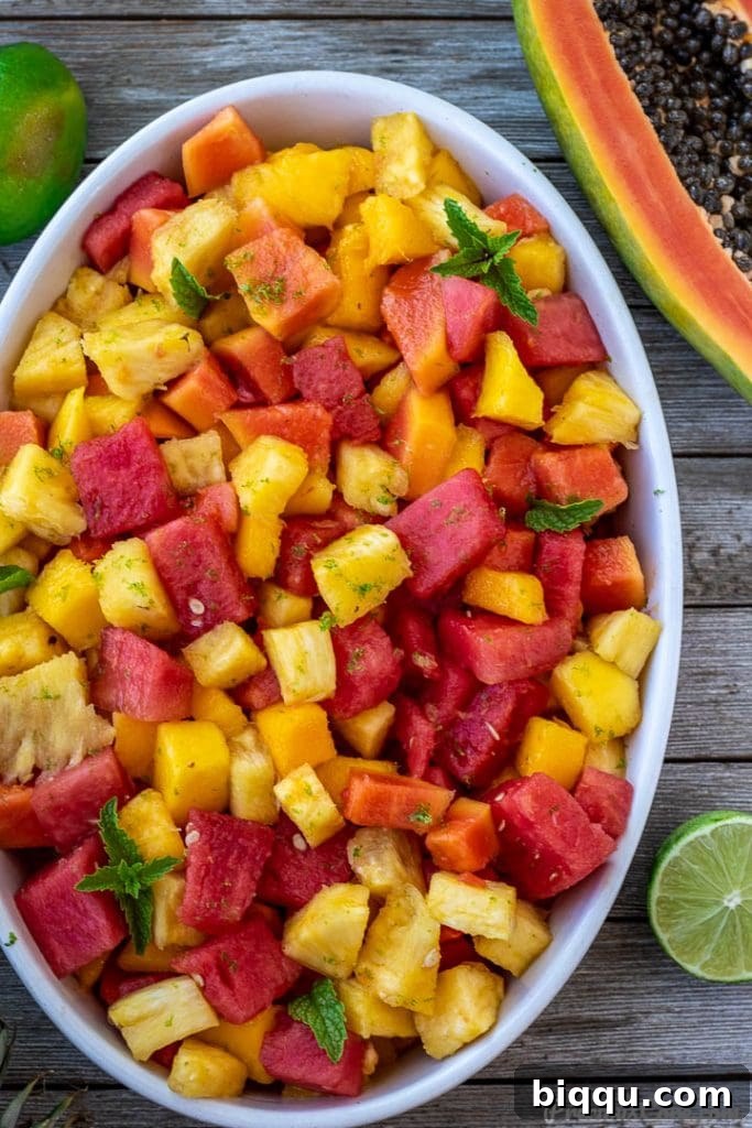 A bowl of fresh tropical fruit salad with half a papaya and a lime on a rustic wooden surface.