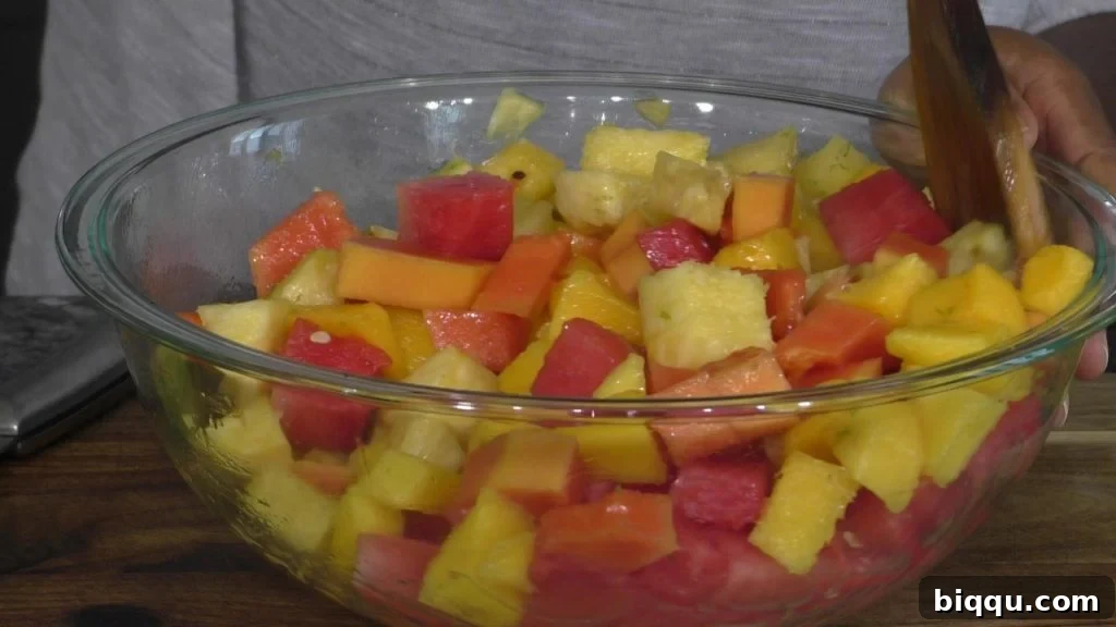 A hand gently mixing a colorful tropical fruit salad in a large mixing bowl with a wooden spoon, showcasing the fresh ingredients.