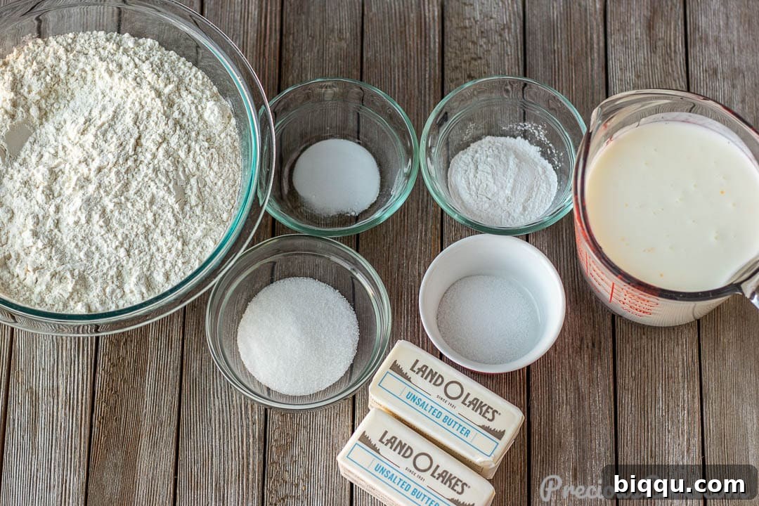 Ingredients for homemade buttermilk biscuits laid out on a table: butter, flour, baking powder, baking soda, salt, sugar, and buttermilk.