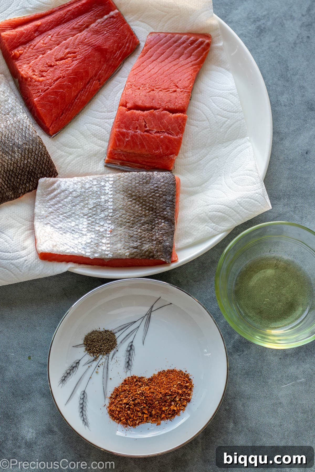 Salmon fillets, a jar of Cajun seasoning, olive oil, and a black pepper grinder on a wooden cutting board.