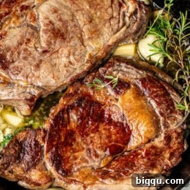 Two ribeye steaks in a cast iron skillet with rosemary, thyme, and garlic.