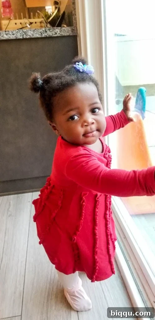 Baby in a pink dress standing at a window, looking at the camera.